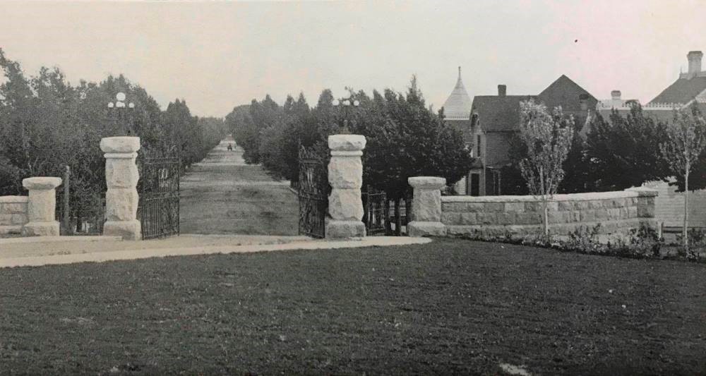 This black and white photo shows a low white wall and pillars between the university and campus.